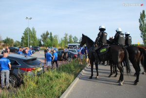 Na zdjęciu widoczna grupa osób na parkingu, obok czterech policjantów na koniach. Policjanci wyposażeni są w kaski i kamizelki.&quot;&gt;