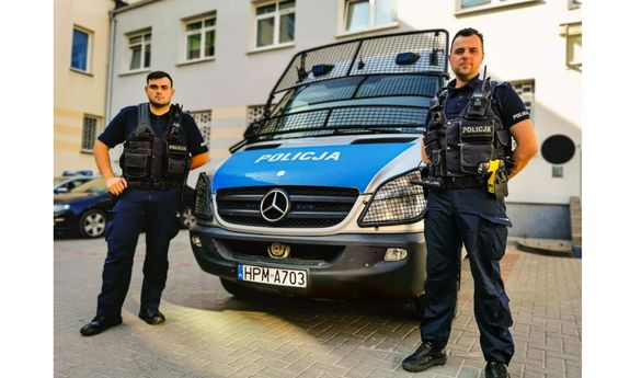 dwaj umundurowani policjanci stoją przy policyjnym radiowozie