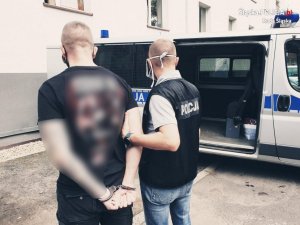 zdjęcie przestawia policjanta kryminalnego z zatrzymanym przy policyjnym radiowozie&quot;&gt;