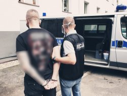 zdjęcie przestawia policjanta kryminalnego z zatrzymanym przy policyjnym radiowozie&quot;&gt;