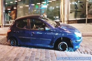 Zdjęcie przedstawia zniszczone niebieskie auto bez dwóch opon na Wrocławskim rynku.