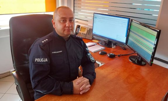 policjant w mundurze siedzi w fotelu przy biurku, z boku widać dwa monitory komputera i telefon