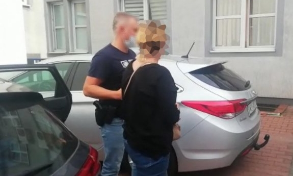 policjant prowadzi zatrzymanego mężczyznę, s w tle samochód na parkingu