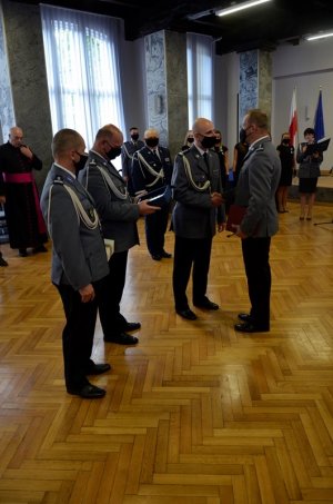 grupa policjantów stojąca na środku pomieszczenia, jeden z nich składa gratulacje drugiemu, pod ścianami stoją goście uroczystości