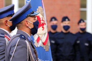 Zdjęcie przedstawia policjantów z pocztu sztandarowego, w tle nowo przyjęci policjanci stojący w szeregu.