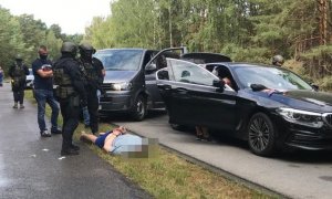 grupa policyjnych kontrterrorystów z leżącym na ziemi zatrzymanym, z tyłu policyjne furgony