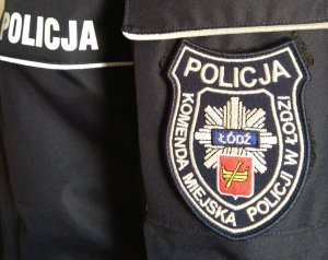 Na mundurze napis POLICJA oraz naszywka z napisem Komenda Miejska Policji w Łodzi a pośrodku naszywki znajduje się odznaka policyjna z herbem Łodzi