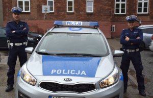 Dwaj policjanci stoją przy radiowozie na dziedzińcu komendy miejskiej Policji