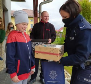 Przed domem umundurowana policjantka wręcza chłopcu tort urodzinowy, z tyłu stoi mężczyzna oraz młoda kobieta, która robi zdjęcia telefonem komórkowym