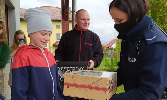 Przed domem umundurowana policjantka wręcza chłopcu tort urodzinowy, z tyłu stoi mężczyzna oraz młoda kobieta, która robi zdjęcia telefonem komórkowym