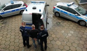 dwaj policjanci stoją z zatrzymanym przed policyjnym furgonem, z tyłu widać inne radiowozy