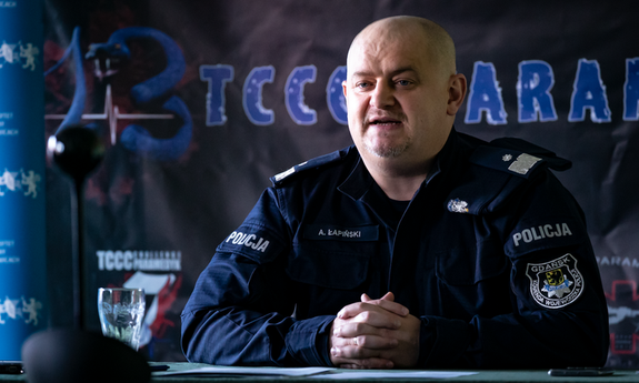 policjant siedzi przy stole, w tle kolorowe banery, na stole stoi szklanka