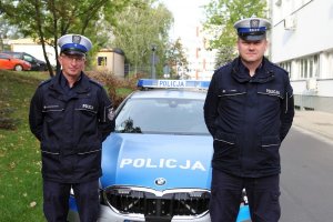 mł. asp. Daniel Szustakowski i asp. szt. Ireneusz Turek pozują do zdjęcia w mundurach na tle oznakowanego bmw