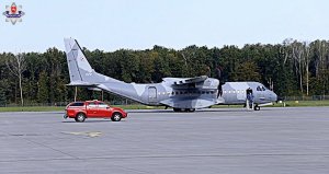 Samolot stoi na płycie lotnisko, wychodzą z niego dwie osoby, po lewej stronie stoi samochód