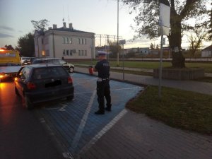 umundurowany policjant w białej czapce  stoi obok zaparkowanego na miejscu dla niepełnosprawnych vw , który za chwile będzie wciągany na widoczną przed nim lawetę