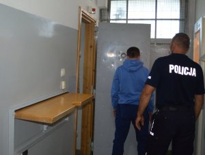 Zatrzymany w niebieskiej bluzie stoi twarzą do drzwi pomieszczenia dla osób zatrzymanych a obok tyłem stoi policjant