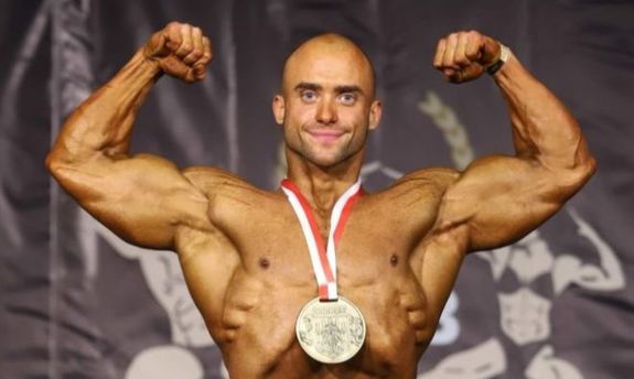 Sierżant sztabowy Marcin Dziadczyk stojący przodem, prezentujący bicepsy, na szyji ma medal