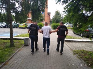 Dwóch policjantów (po bokach) prowadzi jednego z podejrzanych w kajdankach na rękach i na nogach (w środku) - widać ich tyłem, w tle budynek i parking z samochodami