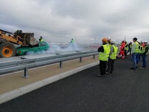 Koparka i samochód, które się zderzyły, dwaj policjanci na miejscu zdarzenia, ludzie w odblaskowych kamizelkach przyglądający się zdarzeniu