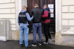 Dwaj policjanci (po bokach) prowadzą drugiego z zatrzymanych (z kajdankami z przodu na rękach, w środku) do jakiegoś budynku, stoją przed drzwiami