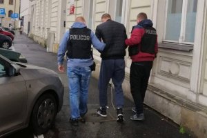 Dwaj policjanci (po bokach) prowadzą drugiego z zatrzymanych (z kajdankami z przodu na rękach, w środku), idą ulicą