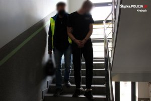 policjant sprowadza po schodach zatrzymanego, który ma założone na ręce i nogi kajdanki zespolone