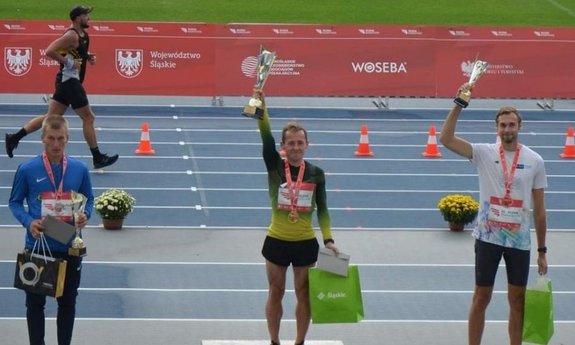 Zawodnicy na podium z medalami i pucharami, za nimi biegnący zawodnik