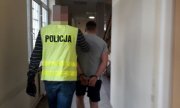 Na zdjęciu zatrzymany mężczyzna w kajdankach. Jest prowadzony korytarzem w jednostce Policji przez nieumundurowanego policjantka, który ma żółtą kamizelkę z napisem Policja
