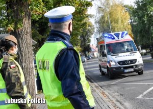 Policjant ruchu drogowego i żołnierka patrzą na ambulans pogotowia ratunkowego.