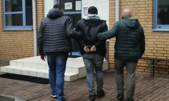dwaj ubrani po cywilnemu policjanci prowadza zatrzymanego mężczyznę do komisariatu Policji