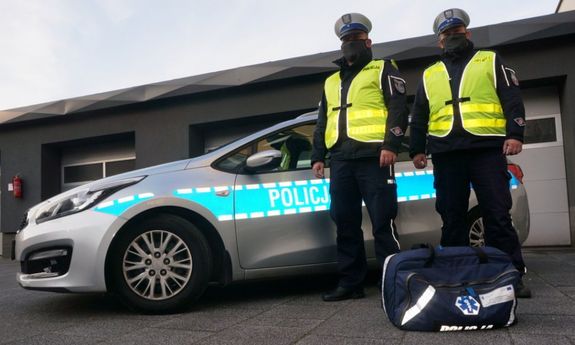 Na zdjęciu widoczni dwaj umundurowani policjanci, stojący na tle oznakowanego radiowozu. Przed nimi na ziemi leży torba medyczna.