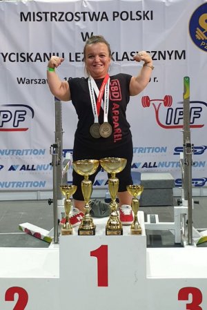 Justyny Kozdryk stojąca na podium (z rękami uniesionymi do góry, medalami na szyi) przed nią stoją puchary