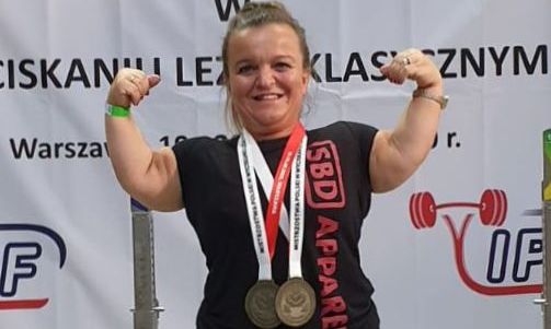 Justyny Kozdryk stojąca na podium (z rękami uniesionymi do góry, medalami na szyi) przed nią stoją puchary