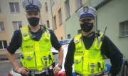 dwaj policjanci ruchu drogowego ubrani w żółte odblaskowe kamizelki z napisem: Policja , stoją przed policyjnym radiowozem