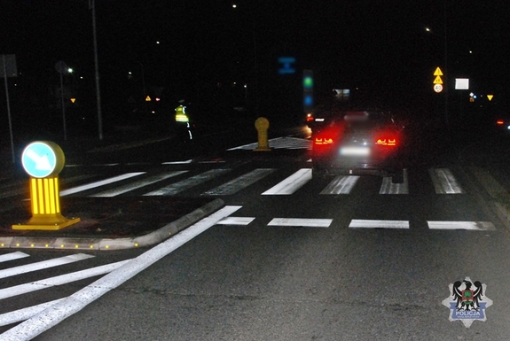 Na zdjęciu nocna porą, przedstawione jest miejsce wypadku drogowego, w tym samochód, który potrącił pieszego oraz policjanta na miejscu zdarzenia nieopodal przejścia dla pieszych