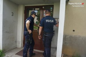 Na fotografii widać dwóch policjantów, którzy stoją przed wejściem do klatki schodowej bloku. Jeden z policjantów dzwoni na domofon