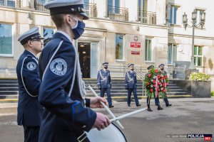 Zdjęcie przed budynkiem Komendy Głównej Policji. Na pierwszym planie dwóch policjantów z Orkiestry Reprezentacyjnej Policji. W tle Komendant Główny Policji wraz Zastępcami oraz policjantka niosąca wieniec.