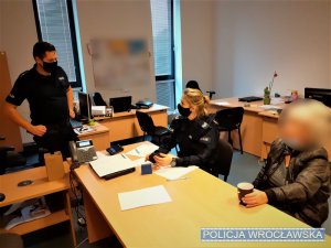 Na zdjęciu biuro na komendzie policji, w którym znajdują się policjant i policjantka oraz kobieta, której pomogła policja