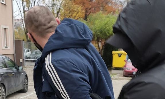 Policjant po cywilnemu prowadzi zatrzymanego. Policjant ma założony na głowę kaptur