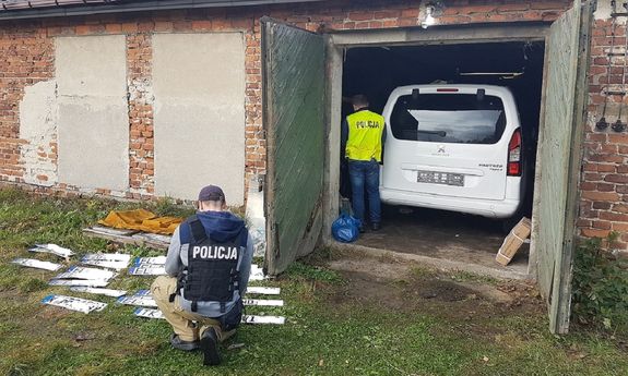 widoczny tył białego pojazdu i stojący obok funkcjonariusz w żółtej kamizelce, przed budynkiem na trawie leżą rozłożone tablice rejestracyjne przy których kuca funkcjonariusz w czarnej kamizelce z napisem policja