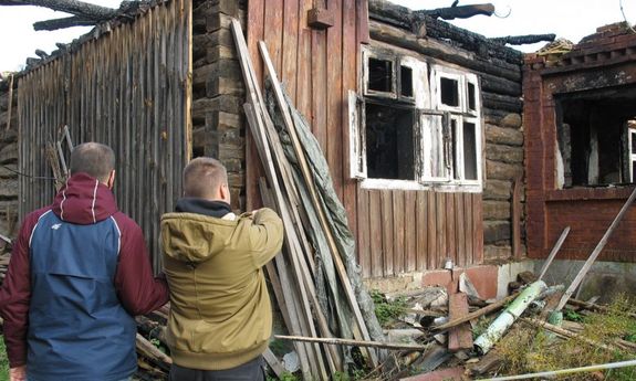 Na zdjęciu widać dwie osoby po lewej stronie policjant w ubiorze cywilnym  granatowo-bordowa kurtka) po prawej stronie podejrzany w kurtce w kolorze khaki. Mężczyźni stoją przed spalonym pomieszczeniem gospodarczym