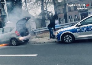 palący się samochód z otwartym bagażnikiem, za nim stoi radiowóz policyjny