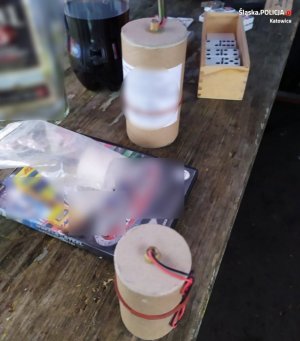 zdjęcie kolorowe: na drewnianym stole ogrodowym ustawione są  3 materiały pirotechniczne, gra komputerowa, butelka po alkoholu i drewniane pudełko z grą domino