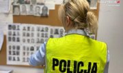 policjantka w kamizelce odblaskowej stoi przodem do tablicy z wizerunkami osób poszukiwanych