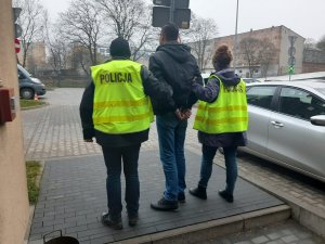 Zdjęcie jest zrobione na dziedzińcu Komendy Miejskiej Policji w Łodzi, widzimy na nim zatrzymanego ubranego w ciemną kurtkę, dżinsy. Jest wysoki, szczupłej budowy ciała, ma ciemne włosy. Ręce ma skute kajdankami. Po jego lewej stronie trzyma go policjant ubrany w żółtą kamizelkę policyjna, a po prawej stronie stoi policjantka, ubrana również w żółtą kamizelkę policyjną&quot;&gt;
