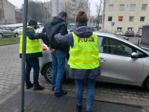 Na zdjęciu widzimy jak sprawca jest wprowadzany przez policjantów do szarego samochodu osobowego. Policjant wraz z policjantką ubrani są w żółte kamizelki policyjne.&quot;&gt;