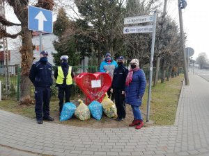 Na zdjęciu widać trzech umundurowanych policjantów oraz kobietę i mężczyznę, którzy stoją przy czerwonym pojemniku w kształcie serca, obok stoją cztery worki foliowe z plastikowymi nakrętkami. W tle widać budynek, ogrodzenie, znaki drogowe, drzewa i krzewy