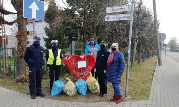 Na zdjęciu widać trzech umundurowanych policjantów oraz kobietę i mężczyznę, którzy stoją przy czerwonym pojemniku w kształcie serca, obok stoją cztery worki foliowe z plastikowymi nakrętkami. W tle widać budynek, ogrodzenie, znaki drogowe, drzewa i krzewy