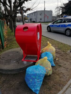 Na zdjęciu widać pojemnik w kształcie serca koloru czerwonego, przy którym stoją cztery worki foliowe z nakrętkami plastikowymi. W tle widać radiowóz, budynek, ogrodzenie, znaki drogowe, samochód, drzewa i krzewy