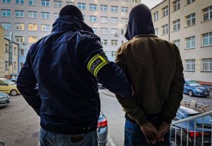 Policjant kryminalny z żółtą, odblaskową opaską na ramieniu z napisem policja. Policjant trzyma zatrzymanego mężczyznę, który ma założone kajdanki na ręce trzymane z tyłu